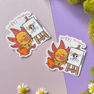 May include: Two stickers featuring a cartoon character with orange hair and a yellow outfit, holding a paintbrush and smiling. The stickers have the text "happy accidents" and a drawing of a skull and crossbones on an easel.