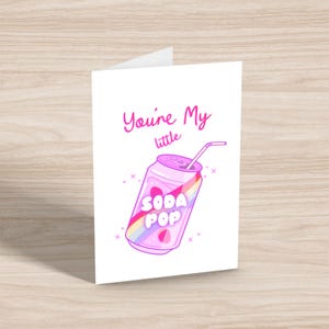 May include: A folded white greeting card with the text "You're My little" in pink script. The card features a cartoon illustration of a pink soda can with a rainbow design and the words "SODA POP". A white straw extends from the can.