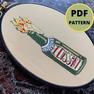 Puede incluir: Arte de bordado en forma de aro que presenta una botella verde con llamas, un cuello blanco y la palabra "DISSENT" en letras rojas y doradas. El aro es negro y el fondo es beige.