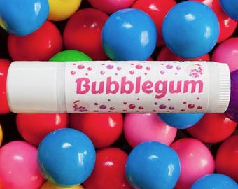 May include: A white cylindrical lip balm tube with a label featuring the word Bubblegum in pink text, surrounded by pink and purple bubble graphics, positioned over a background of many colorful, shiny, round gumballs in red, blue, yellow, and green.