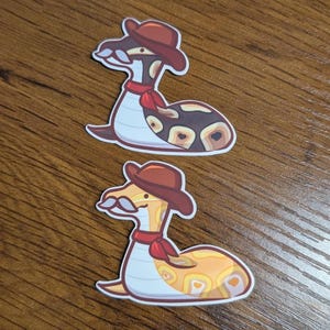 Puede incluir: Dos pegatinas con dibujos de serpientes con sombreros de vaquero y pañuelos rojos. Una serpiente es marrón y beige con un patrón de manchas, la otra es amarilla y naranja con bigote. Sobre una superficie de madera.