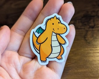 Könnte beinhalten: Ein Aufkleber mit einem orangefarbenen Cartoon-Drachen mit einem türkisfarbenen Flügel. Der Drache hat einen weißen Bauch und einen weißen Streifen auf dem Rücken.