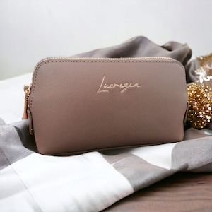Può includere: Una pochette con cerniera di colore marrone chiaro con la parola "Lucenza" ricamata in oro sul davanti.