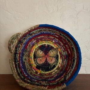 May include: A circular trivet made from fabric strips in a spiral pattern. The trivet features a butterfly applique in the centre and is made from various coloured fabrics, including blue, red, green, and yellow. Designed to protect surfaces from heat.