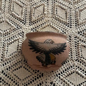 May include: A small, round, light brown pottery bowl with a hand-painted eagle design. The eagle is depicted with outstretched wings in black, white, and yellow. The bowl is likely handmade and has a rustic, artisanal aesthetic.