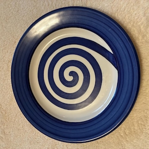 May include: A round ceramic plate with a blue rim and a white and blue spiral design in the center. The plate has a glossy finish and is suitable for serving food. The plate is a decorative item.