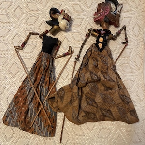 May include: Two Indonesian Wayang Golek puppets. The puppets have painted faces, ornate headdresses, and detailed fabric costumes in shades of brown, black, and gold. Each puppet is held with wooden rods.