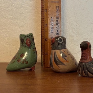 May include: Three small, hand-painted ceramic bird figurines. The owl is green with red and brown accents. The other two birds are tan, black, and red. A wooden ruler with the text "RAZOR SHARPENING" is behind the figurines.