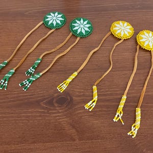 May include: Handmade beaded hair accessories. The image shows several hair ties with circular beaded designs in green and white, and yellow and white. The ties have long, thin leather straps and beaded tassels at the ends.