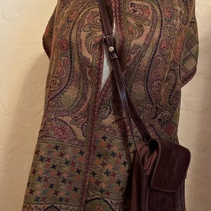 May include: A long, rectangular scarf featuring a multi-colored paisley pattern in shades of brown, green, and orange, finished with fringe at the bottom. A dark brown crossbody bag with a long, adjustable strap hangs over the scarf.