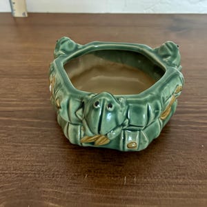 May include: A green ceramic frog-shaped planter with a rectangular opening. The planter has a glossy finish and is decorated with frog faces and leaf details. The interior is a light tan color.