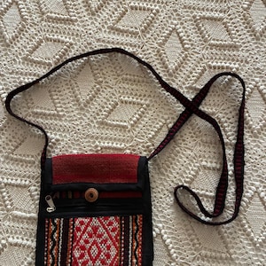 May include: A black crossbody bag featuring a red woven flap, a front zippered pocket, and a central wooden button closure. The bag includes a long, thin, black and red patterned shoulder strap and a rectangular front panel with red, orange, and white geometric woven designs.