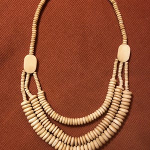 May include: A three-strand necklace made of natural beige wood beads. The necklace has a simple clasp closure.