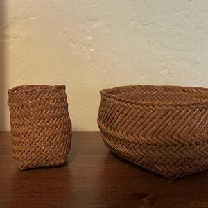 May include: Two small brown woven baskets with a herringbone pattern. The taller basket is approximately 2 inches high, and the wider, shorter basket is approximately 1.5 inches high, as indicated by the wooden ruler placed vertically beside them.