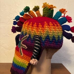 May include: A multi-colored crochet hat featuring a vibrant rainbow gradient pattern. The top is adorned with several small, upright, carrot-shaped yarn protrusions in various colors. A long, rainbow-striped earflap extends downward.