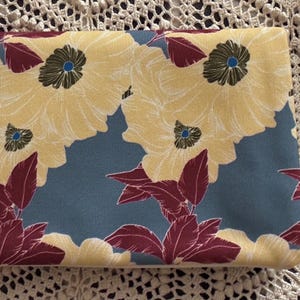 May include: A rectangular fabric pouch with a floral pattern. The design features large yellow flowers with dark centers and maroon leaves against a blue background. The pouch is on a white lace surface.