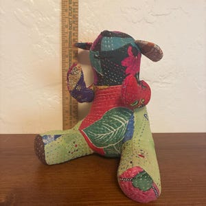 May include: A stuffed animal toy made from patchwork fabric with floral and leaf patterns in shades of green, red, and blue. A wooden ruler placed behind the toy shows it stands approximately 7 inches tall, featuring visible hand-stitched details.