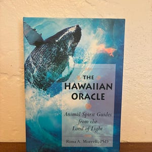 May include: A rectangular book titled The Hawaiian Oracle: Animal Spirit Guides from the Land of Light by Rima A. Morrell, PhD. The cover features a dark blue whale breaching through bright blue ocean waves with white sea foam and spray.