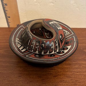 May include: A small, handcrafted ceramic sculpture featuring a coiled, abstract design. The piece is primarily black with red and white accents, set on a matching circular base. The artwork is displayed on a wooden surface.