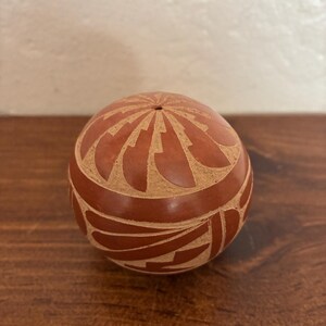 May include: A round, reddish-brown decorative object with a carved geometric design. The object has a small hole at the top and is resting on a wooden surface. The carved design is a lighter tan color, creating a contrast with the darker base color.