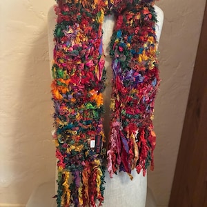 May include: A multi-coloured scarf featuring a textured, shaggy knit construction with various fabric ribbons in shades of red, orange, yellow, green, blue, and purple. The scarf includes a small black tag with the text Rising Tide in white.