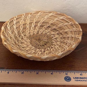 May include: A handwoven, oval-shaped basket made of natural-colored straw. The basket features a spiral design in the center and a woven rim. The basket is approximately 6 inches in diameter.