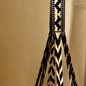 May include: Hanging woven basket with a circular metal ring. The basket features a geometric pattern in white, black, and blue. Braided cords support the basket, and the top strap has a repeating pattern.