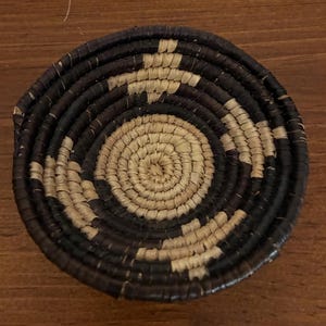 May include: A round, handwoven basket with a spiral design. The basket features alternating bands of dark brown and natural beige fibers. The center of the basket is a light beige color, and the outer edge is dark brown.