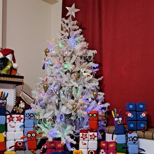 May include: A white Christmas tree decorated with lights and ornaments, surrounded by a group of colorful plush toys shaped like blocks with faces. The toys are arranged in a semi-circle around the tree.
