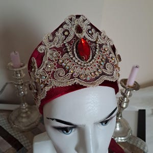 Peut inclure: Un couvre-chef en velours bordeaux orné de dentelle dorée et de cristaux étincelants. Une grosse pierre précieuse rouge est centrée sur le dessus. Le couvre-chef est présenté sur une tête de mannequin, mettant en valeur son design orné.