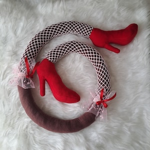 Puede incluir: Un objeto decorativo de felpa con forma de piernas, con medias de rejilla marrones y zapatos de tacón rojos. La base es marrón, con encaje rosa y cintas rojas. Diseño lúdico y único.