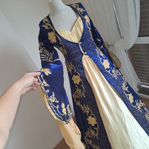 Könnte beinhalten: Ein langes, fließendes Kleid mit einem blauen und goldenen Stickmuster. Das Kleid hat ein tailliertes Oberteil und einen weiten Rock.
