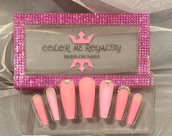 May include: A set of pink press-on nails with a clear base and a gradient of pink to white. The nails are in a clear plastic case with a pink and rhinestone border. The case has the text "Color Me Royalty" and "Press On Nails" printed on it.