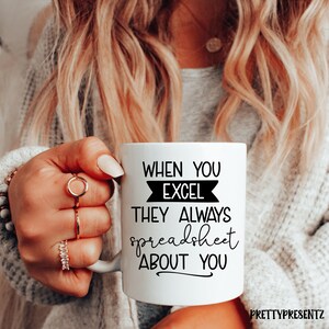 Könnte beinhalten: Weiße Keramiktasse mit schwarzem Text: "WHEN YOU EXCEL THEY ALWAYS spreadsheet ABOUT YOU". Die Tasse wird von einer Person mit blonden Haaren und Ringen gehalten. Die Tasse hat einen Henkel auf der rechten Seite.