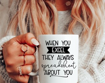 Puede incluir: Taza de cerámica blanca con texto negro: "WHEN YOU EXCEL THEY ALWAYS spreadsheet ABOUT YOU". La taza la sostiene una persona con cabello rubio y anillos. La taza tiene un asa en el lado derecho.