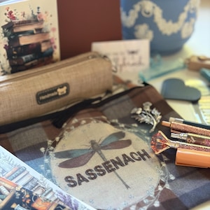 May include: A brown plaid fabric pouch featuring a printed dragonfly and the text SASSENACH, accompanied by a silver thistle brooch, several decorative pens with glitter and gemstone toppers, a beige pencil case, and illustrated stationery items.