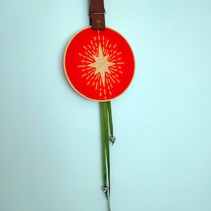 May include: A red embroidery hoop with a yellow starburst design and green ribbon hanging from the bottom.