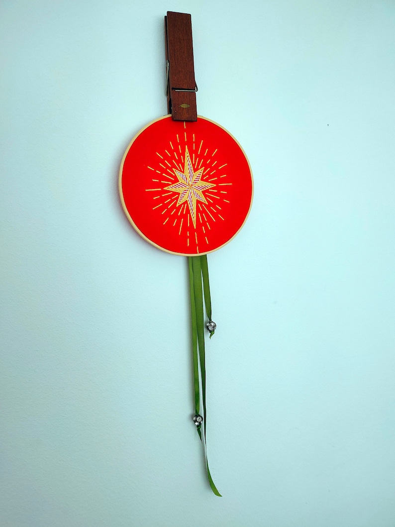 May include: A red embroidery hoop with a yellow starburst design and green ribbon hanging from the bottom.