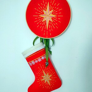 May include: A red Christmas stocking with a white lace trim and a gold star with yellow rays embroidered on it. The stocking is hanging from a red and gold hoop with a similar star design.