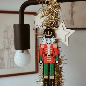 May include: A Christmas ornament of a nutcracker soldier in a red coat with green pants and black boots. The ornament is hanging with gold tinsel and wooden star ornaments. A black lamp and a framed picture are in the background.