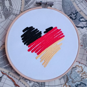 Puede incluir: Dise&ntilde;o bordado en forma de coraz&oacute;n en los colores de la bandera alemana: negro, rojo y dorado. El dise&ntilde;o est&aacute; cosido en tela blanca y montado en un aro de bordado de madera.