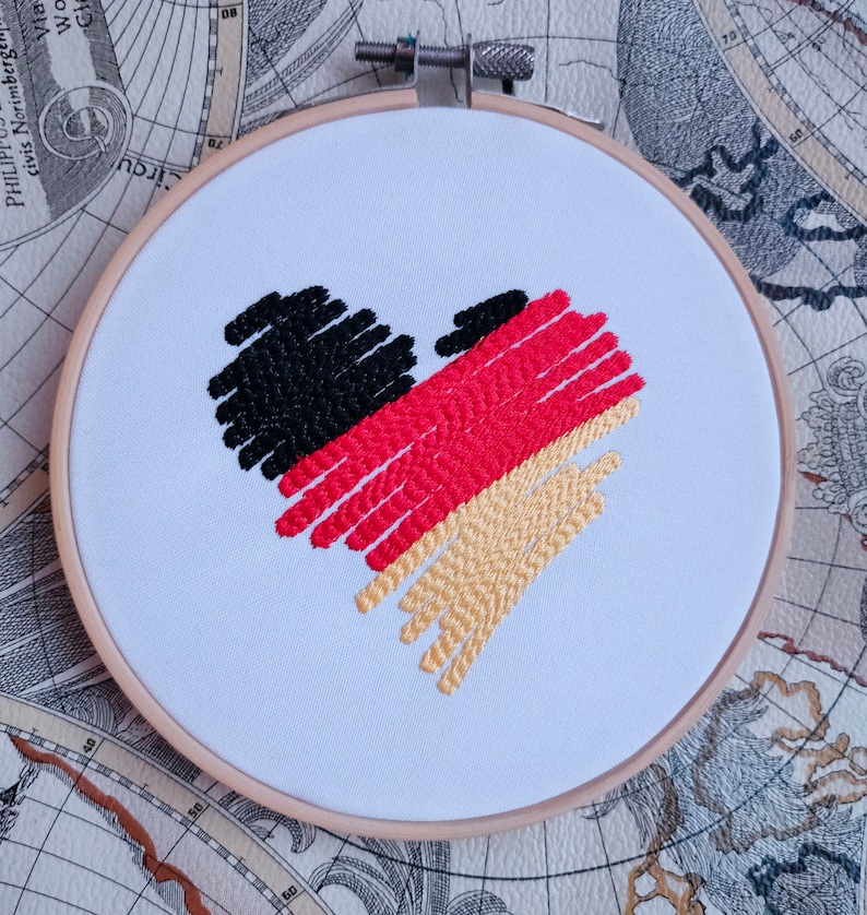 Puede incluir: Dise&ntilde;o bordado en forma de coraz&oacute;n en los colores de la bandera alemana: negro, rojo y dorado. El dise&ntilde;o est&aacute; cosido en tela blanca y montado en un aro de bordado de madera.