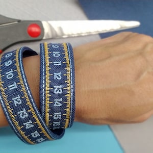 May include: Two blue denim bracelets with gold stitching and white numbers. The bracelets are wrapped around a person's wrist. The numbers on the bracelets are 8, 9, 10, 11, 12, 13, 14, 15, and 16.