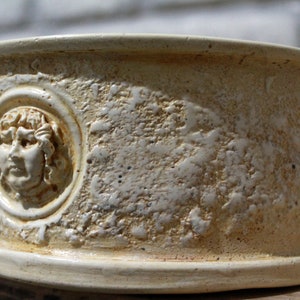 May include: A white ceramic planter with a raised design of a face in a circle. The planter has a rough, textured surface.