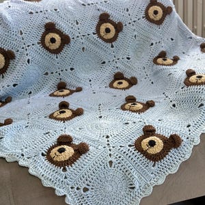 Può includere: Una copertina per neonati in crochet blu chiaro con un motivo di orso marrone. La copertina è realizzata con un punto delicato e presenta un bordo a festoni.
