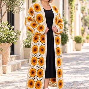 Peut inclure: Un long gilet en crochet ouvert sur le devant, orné d'un motif de tournesols répété dans des tons jaune, orange et brun foncé, bordé de fil crème. Le vêtement possède des manches longues et un ourlet au sol, porté sur une robe noire.