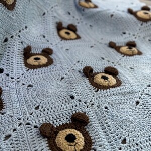 May include: A light blue crochet blanket featuring a repeating pattern of square motifs. Each square contains a centered, three-dimensional bear face applique crafted in dark brown and tan yarn with a small black nose and stitched mouth details.