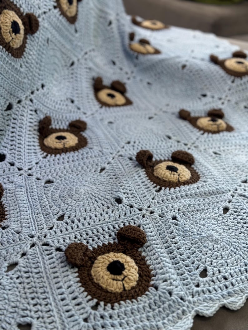 May include: A light blue crochet blanket featuring a repeating pattern of square motifs. Each square contains a centered, three-dimensional bear face applique crafted in dark brown and tan yarn with a small black nose and stitched mouth details.