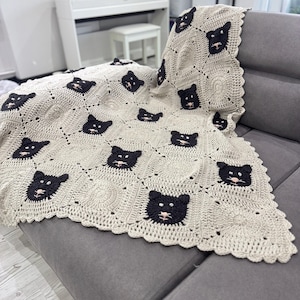May include: A beige crocheted blanket with a repeating pattern of black cat faces and pink noses. The blanket is draped over a grey sofa, highlighting its detailed design and texture. The edges have a scalloped trim.
