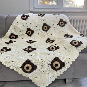 May include: A cream-colored crochet blanket featuring a repeating pattern of square motifs, each decorated with a dark brown bear face. The bear faces consist of a dark brown circular outline with a lighter tan center, small ears, and a black stitched nose.
